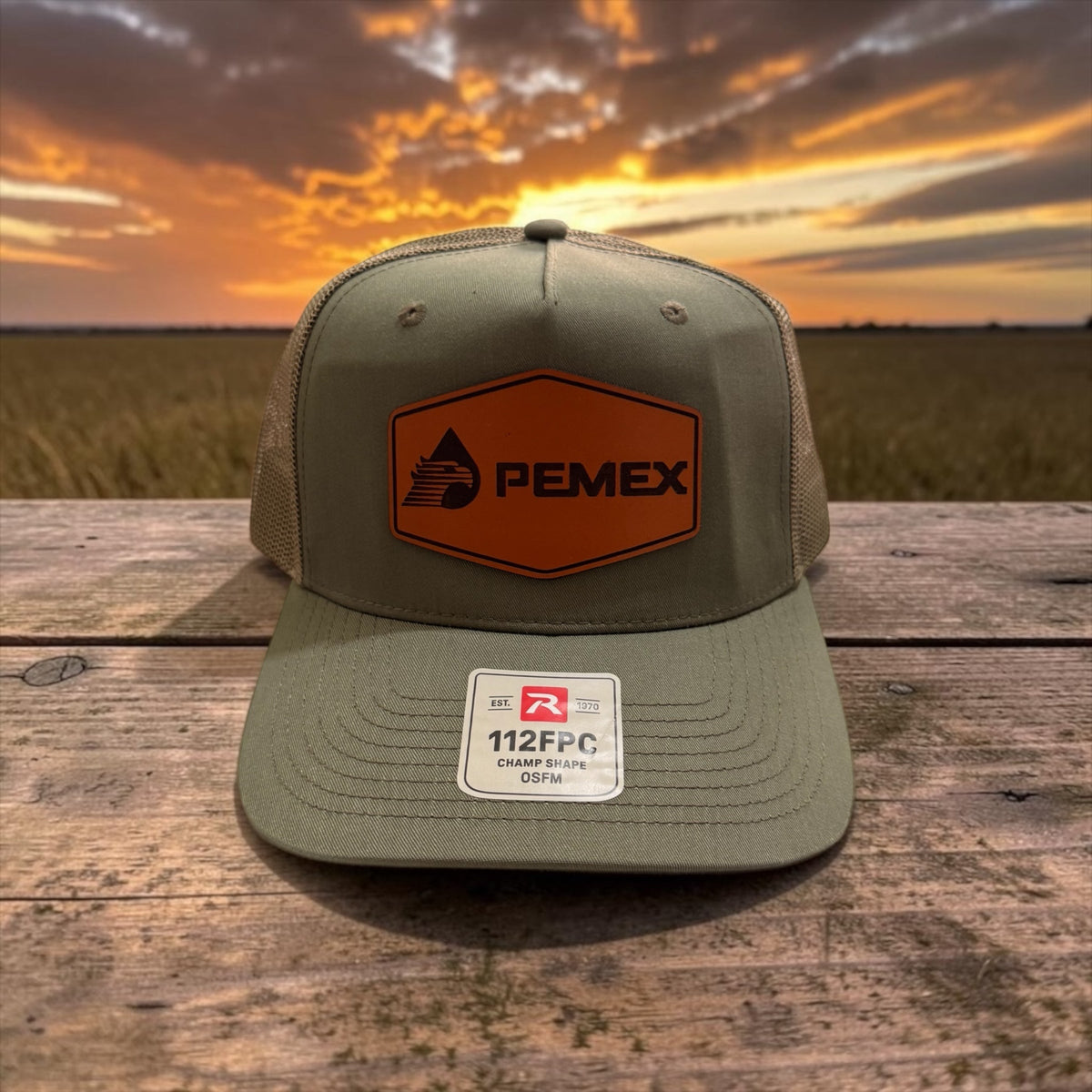 J&G Workwear olive green trucker cap with a PEMEX logo patch and breathable mesh back on a rustic wooden surface.